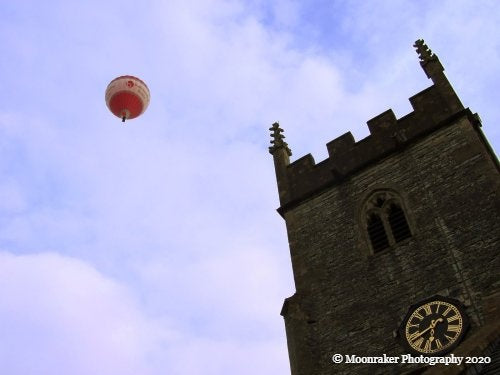 Photograph of a hot air balloon depicted in the air beside the top of a parish.
