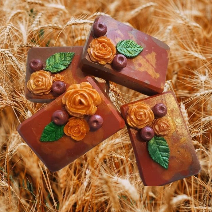 Burnt orange colored soap bare base with a green leaf, two cranberries, and a golden rose on top, all made of soap.