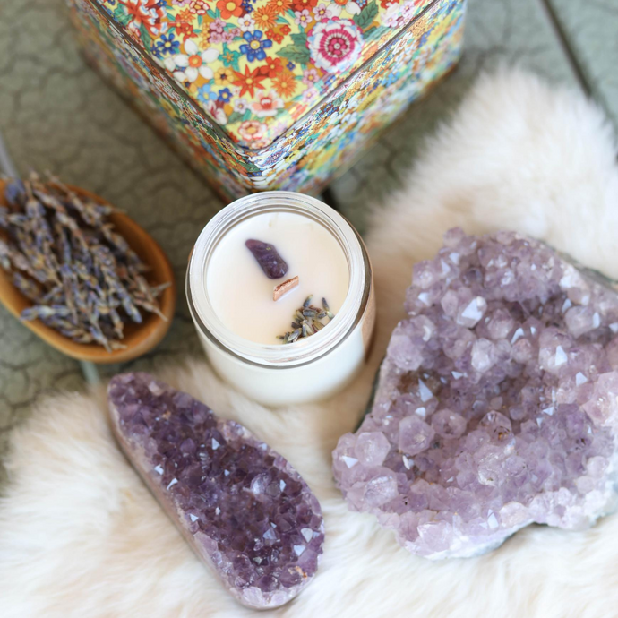 White candle in glass jar with a wooden wick, a purple amethyst crystal, and some dried lavender embedded in the top.