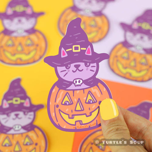 a cute lavender cat has its head poking out of a carved jack-o-lantern, the cat is wearing a black cape with a skull clasp and a black witch hat with a yellow buckle