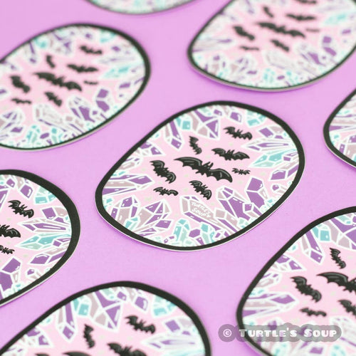 an oval shape sticker with blue and purple multifaceted crystals lining the edges, there are black bats in the center against a pink background