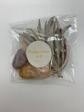Load image into Gallery viewer, The happiness kit, a clear plastic bag containing an orange selenite stick, a white sage sprig, a mini shell, a citrine stone, and lepidolite.
