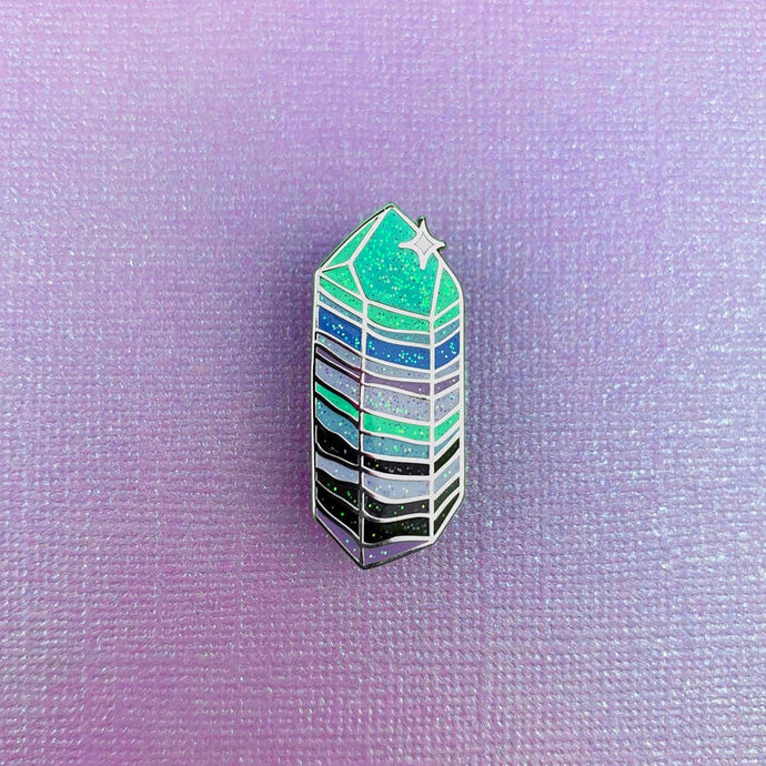 Enamel pin depicting a sparkling, stylized crystal with the colors: turquoise, blue, white, grey, and black.