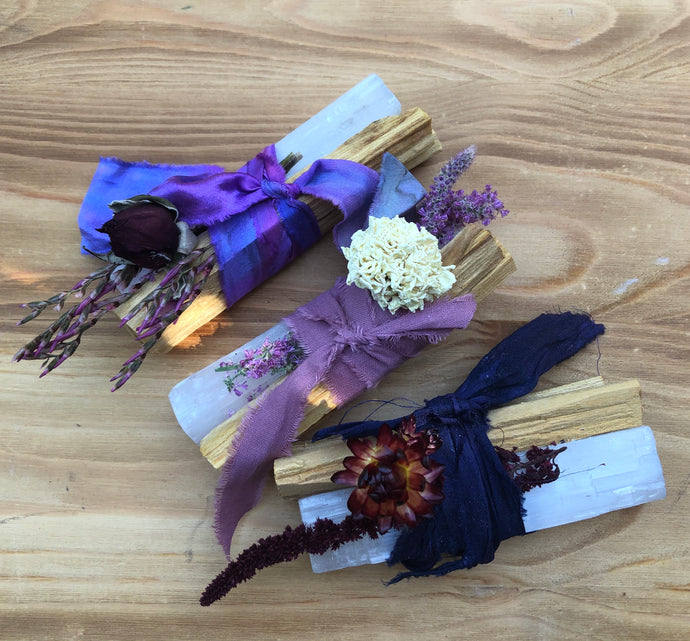 Bundles containing Palo Santo healing wood, a large selenite crystal, and flowers, bound by purple ribbon.