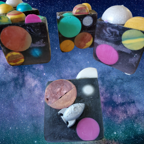 Square, glittery black soaps with colorful planets embedded. One of the soaps has a small silver spaceship adhered to the front.